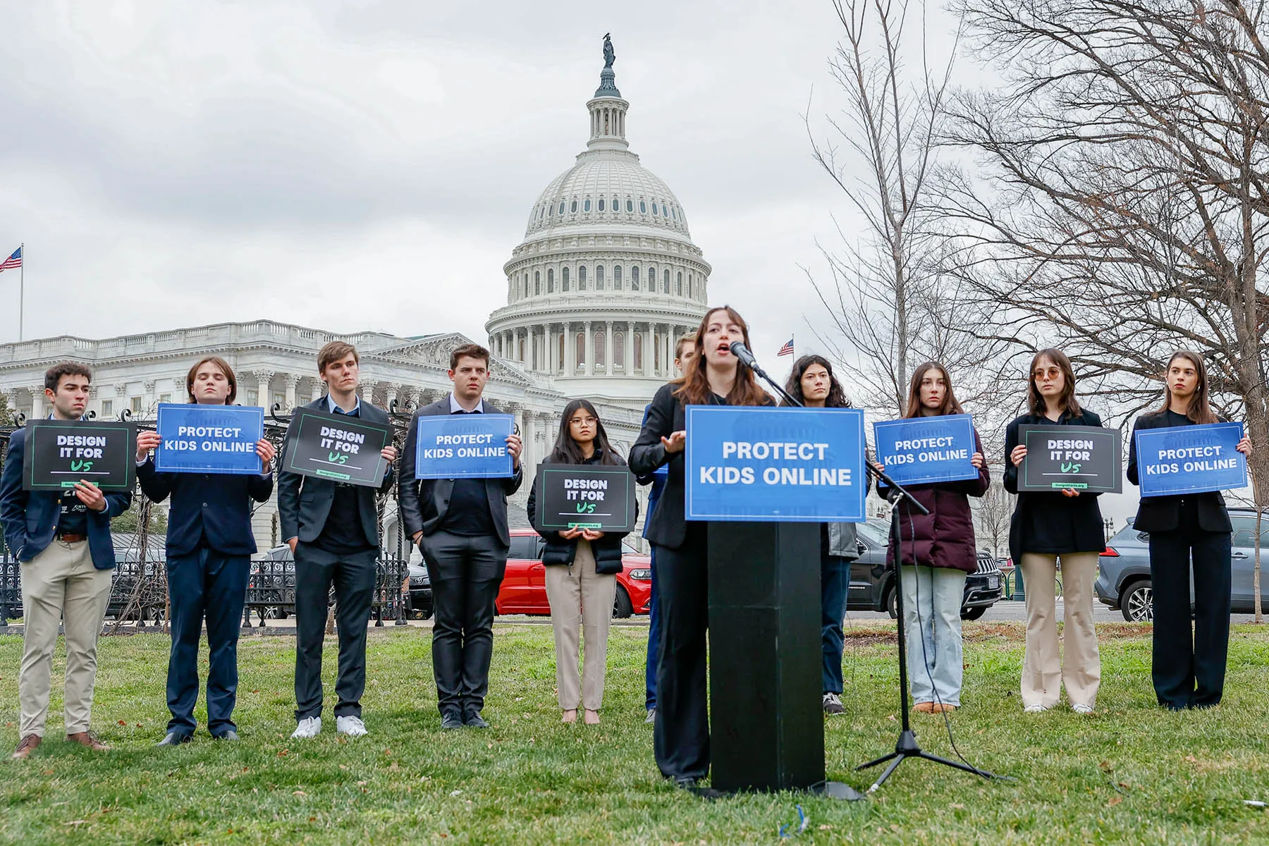 A row of people stand behind a person at a podium in front of the U.S. Capitol, all bearing signs that say Protect Kids Online or Design it For Us.