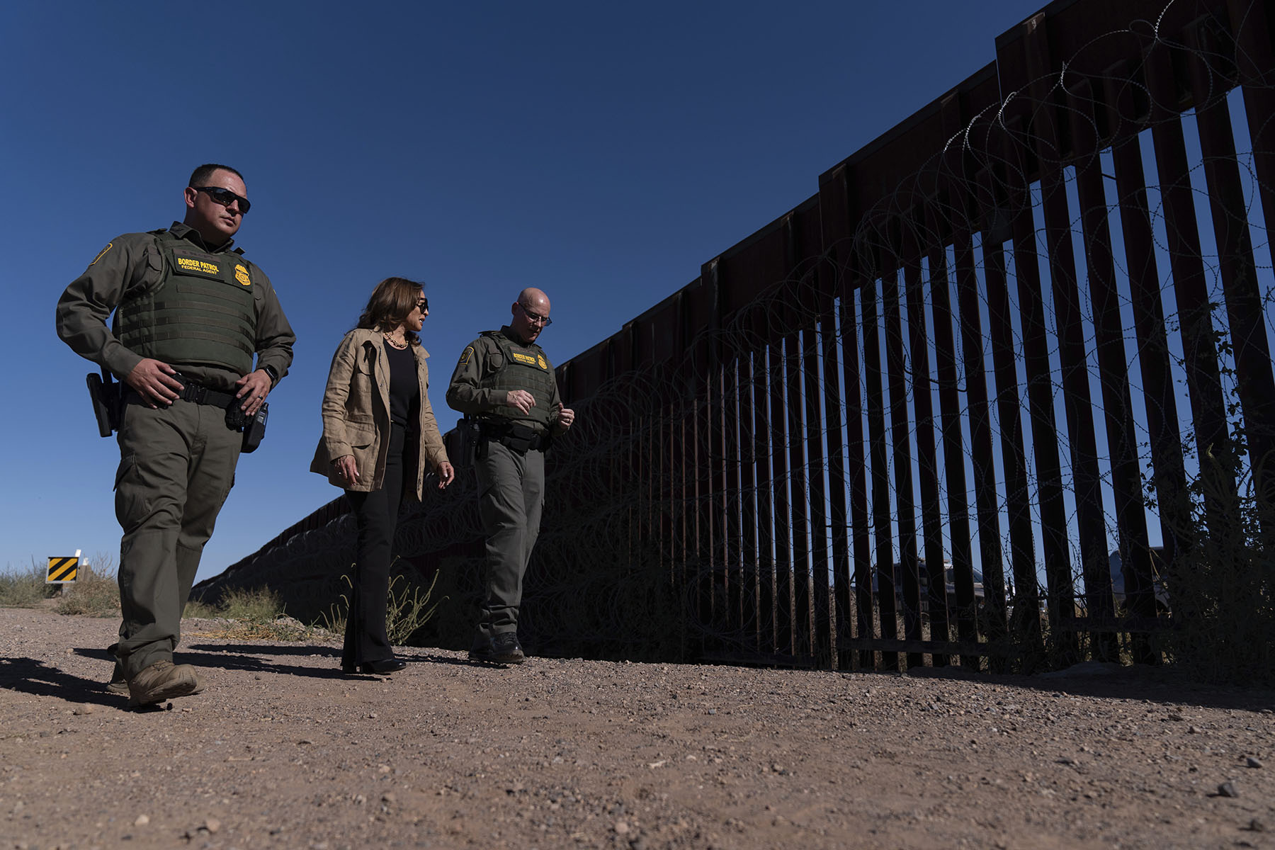 Kamala Harris walks along the U.S.-Mexico border wall with two Border Patrol agents while discussing border security.