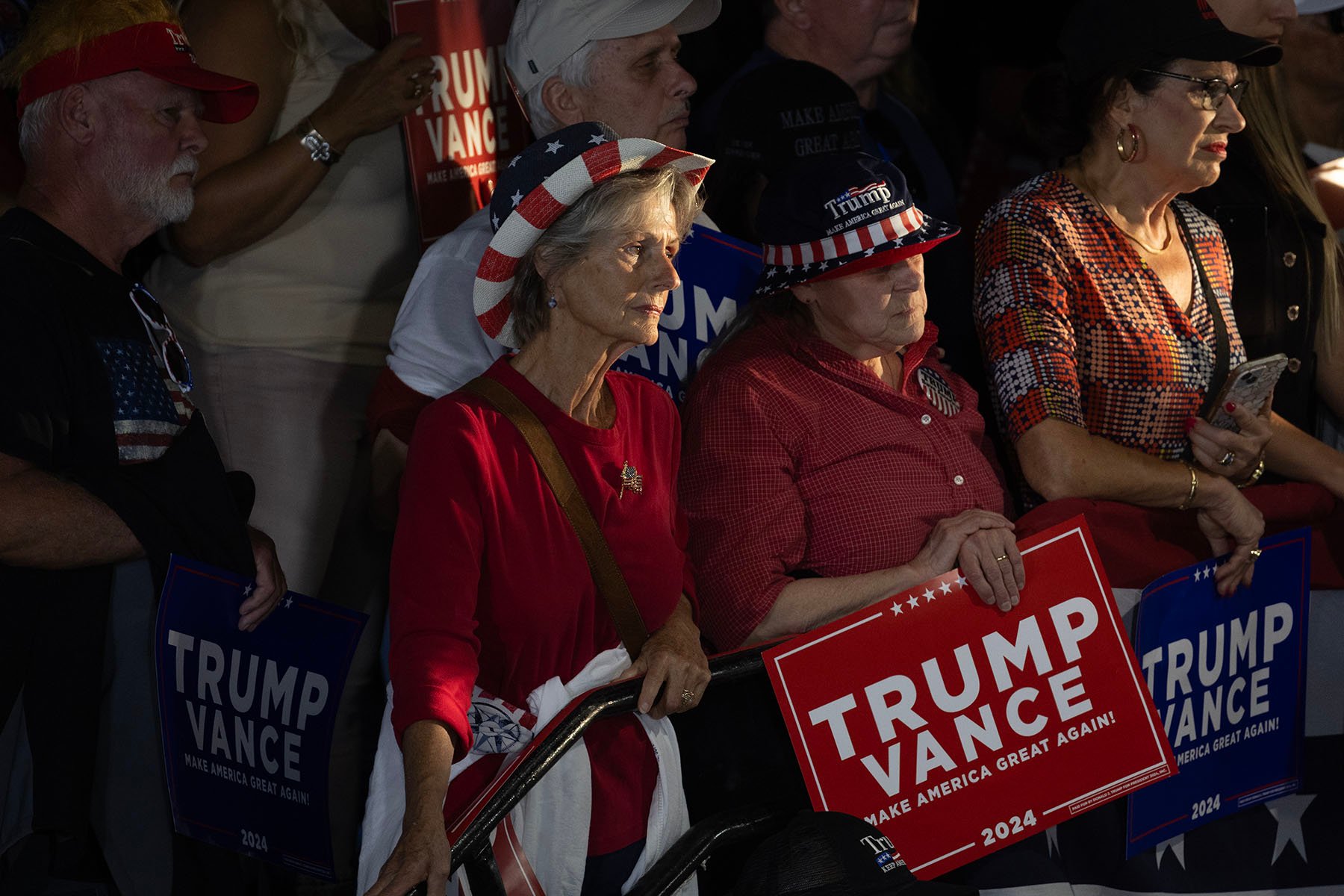 A group of women listen as Sen. JD Vance speaks at a campaign event, some wearing patriotic hats adorned with American flag designs, holding signs that read 'TRUMP VANCE 2024.'