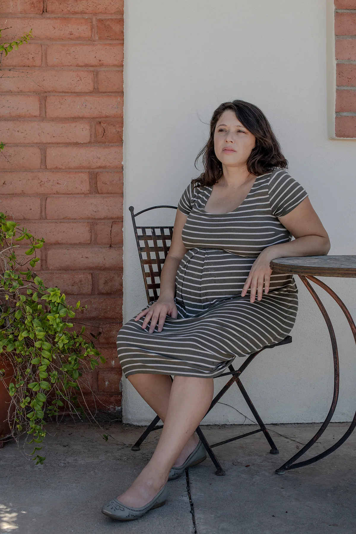 A pregnant woman sits on a metal chair against the backdrop of a white wall and brick exterior, looking into the distance with a thoughtful expression.