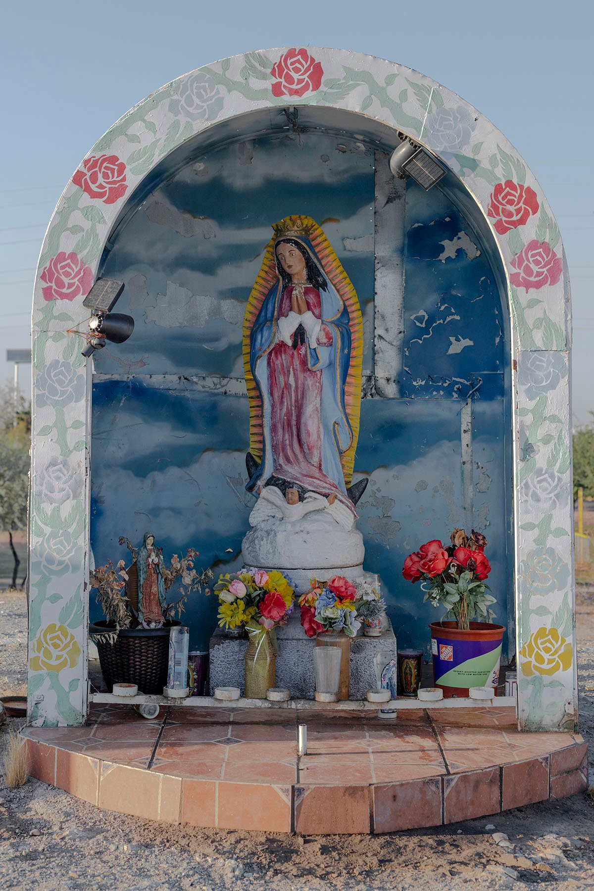 A painted statue of Our Lady of Guadalupe is displayed in a decorative niche surrounded by flowers and candles, set against a blue sky background.