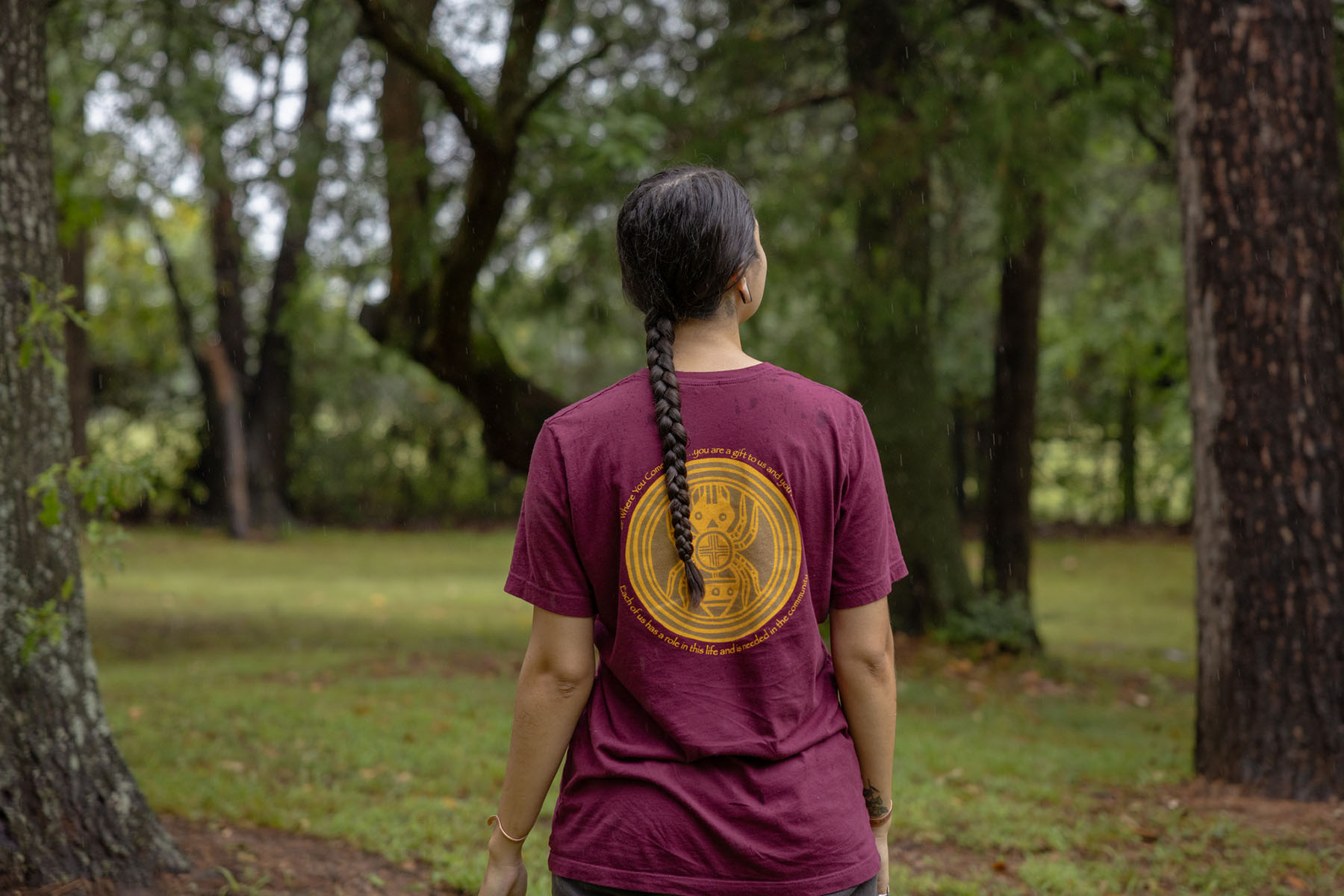 A native woman stands in a forest with her back facing the camera in this anonymized portrait.