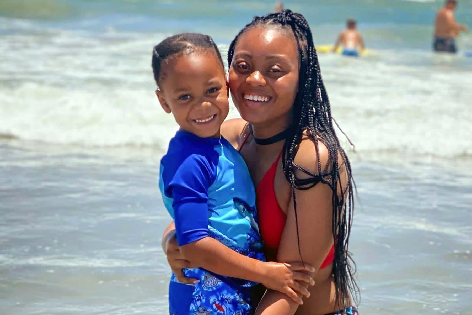 Una sonriente Amber Nicole Thurman en traje de baño rojo sosteniendo a su pequeño hijo en la playa, juntos en el agua, con olas y otros bañistas al fondo.