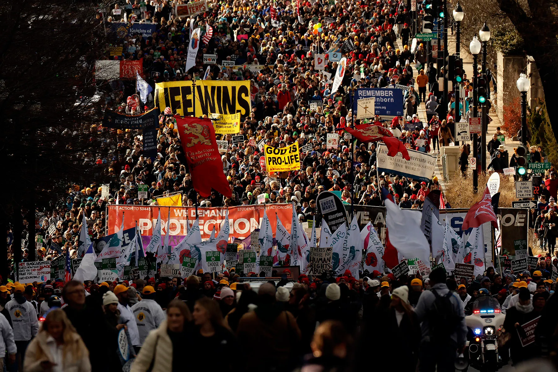 A massive crowd of pro-life activists marches, holding banners and signs with anti-abortion slogans, including "God is Pro-Life" and "We Demand Protection," as they walk through the streets of Washington, D.C.