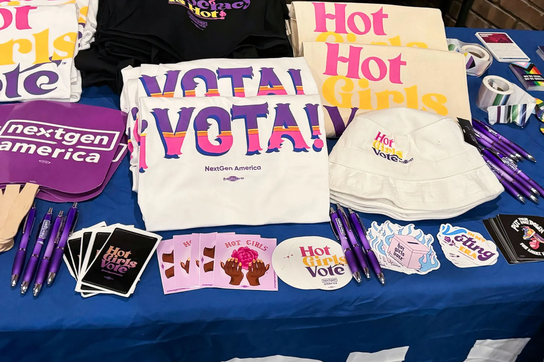 A table offers brightly colored shirts, stickers, pens, bucket hats and fans with slogans "Vota!" and "Hot Girls Vote."