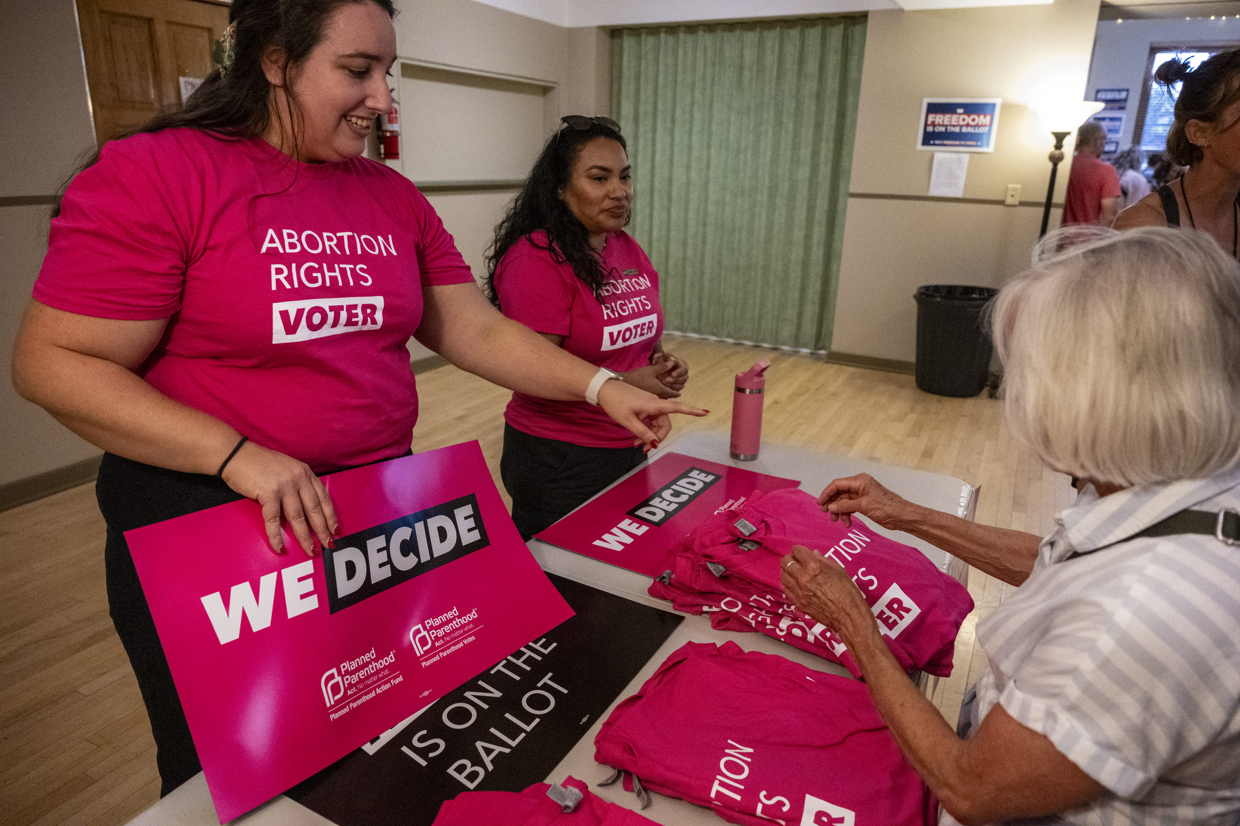 Women wearing pink shirts that say "Abortion Rights Voter" pass out shirts to event attendees at a table.