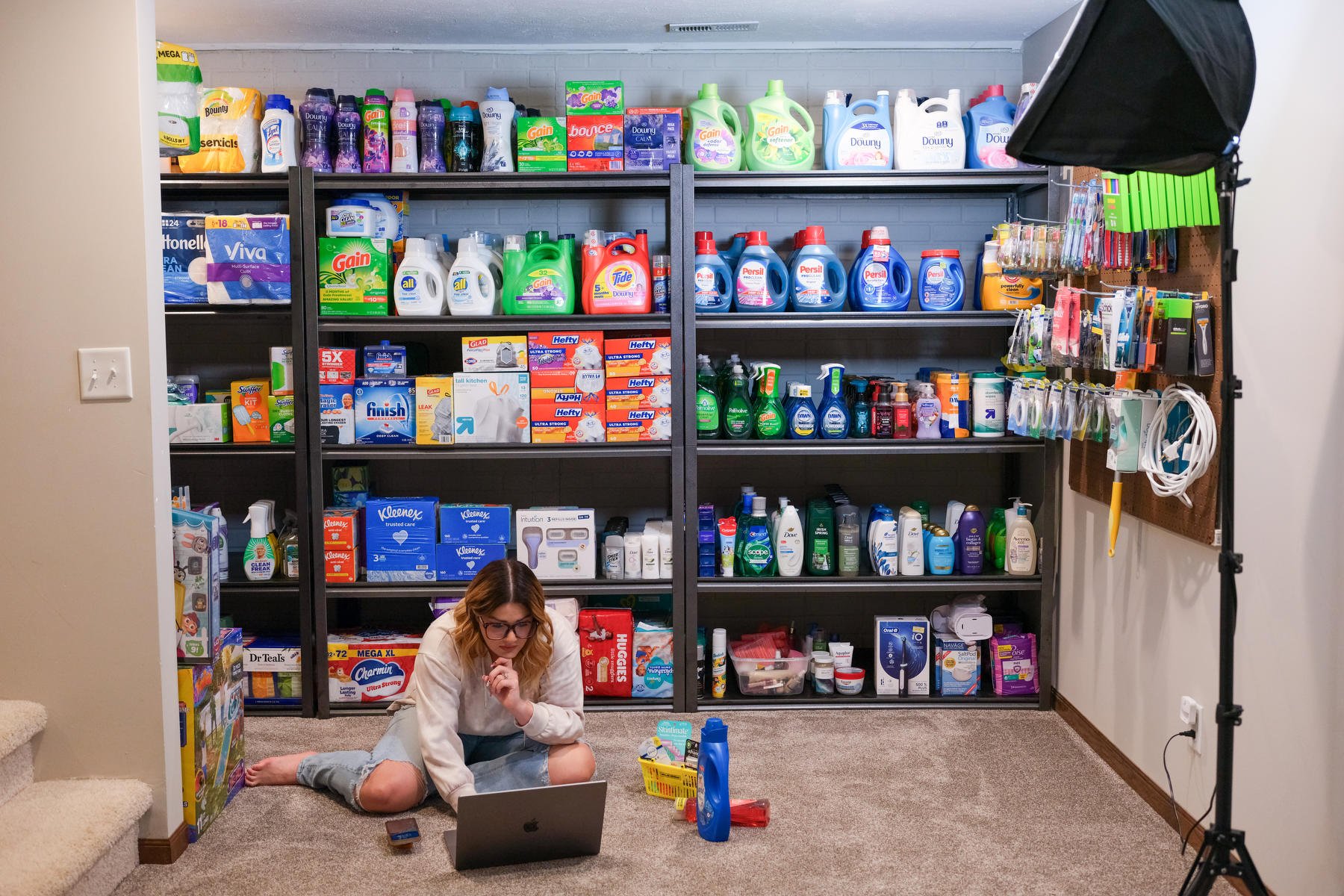 A woman sits on a carpeted floor in a room with a studio light in the foreground and many shelves of organized household products behind her.