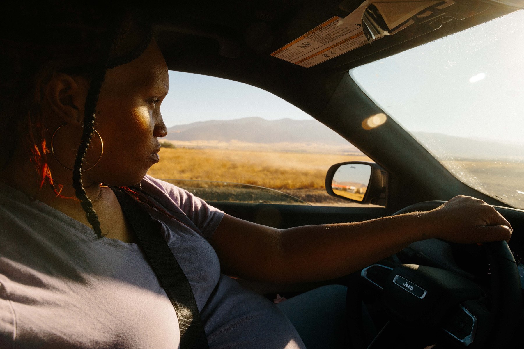 A woman drives a car past a barren landscape with the the sun in her eyes.