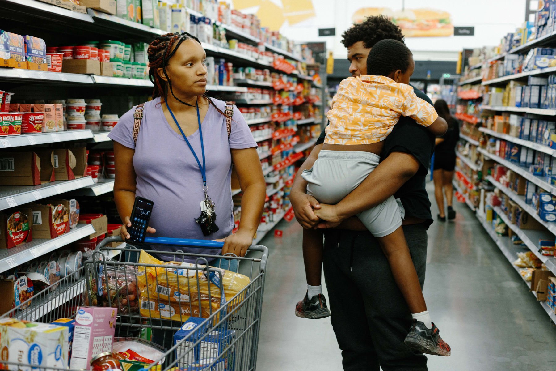 A pregnant woman pushes a grocery cart while walking alongside a young man carrying a small boy.