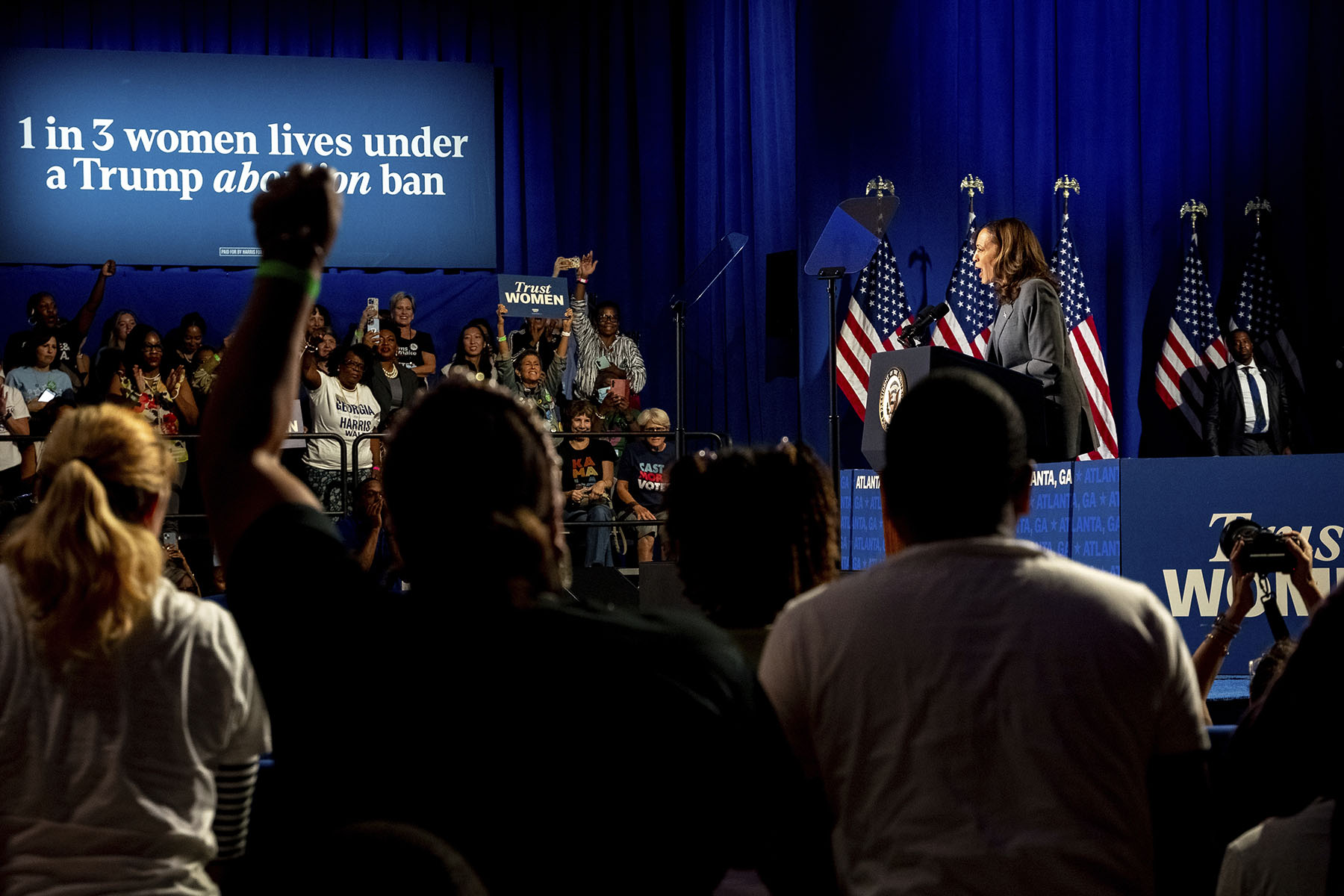 Vice President Kamala Harris speaking at a podium with American flags behind her, addressing a crowd at a campaign rally in Atlanta. A large sign behind her reads, '1 in 3 women lives under a Trump abortion ban.'