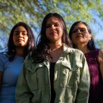 Adriana Grijalva stands with her sister and mother, posing for a portrait outdoors at their home in Tucson, Arizona. All three women look confidently at the camera, with trees in the background.