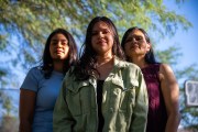 Adriana Grijalva stands with her sister and mother, posing for a portrait outdoors at their home in Tucson, Arizona. All three women look confidently at the camera, with trees in the background.