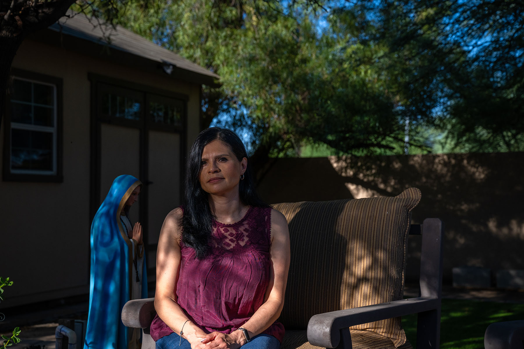 Adriana Grijalva’s mother sits outdoors in their garden at home, looking at the camera with a calm expression. A statue of the Virgin Mary is partially visible in the background.