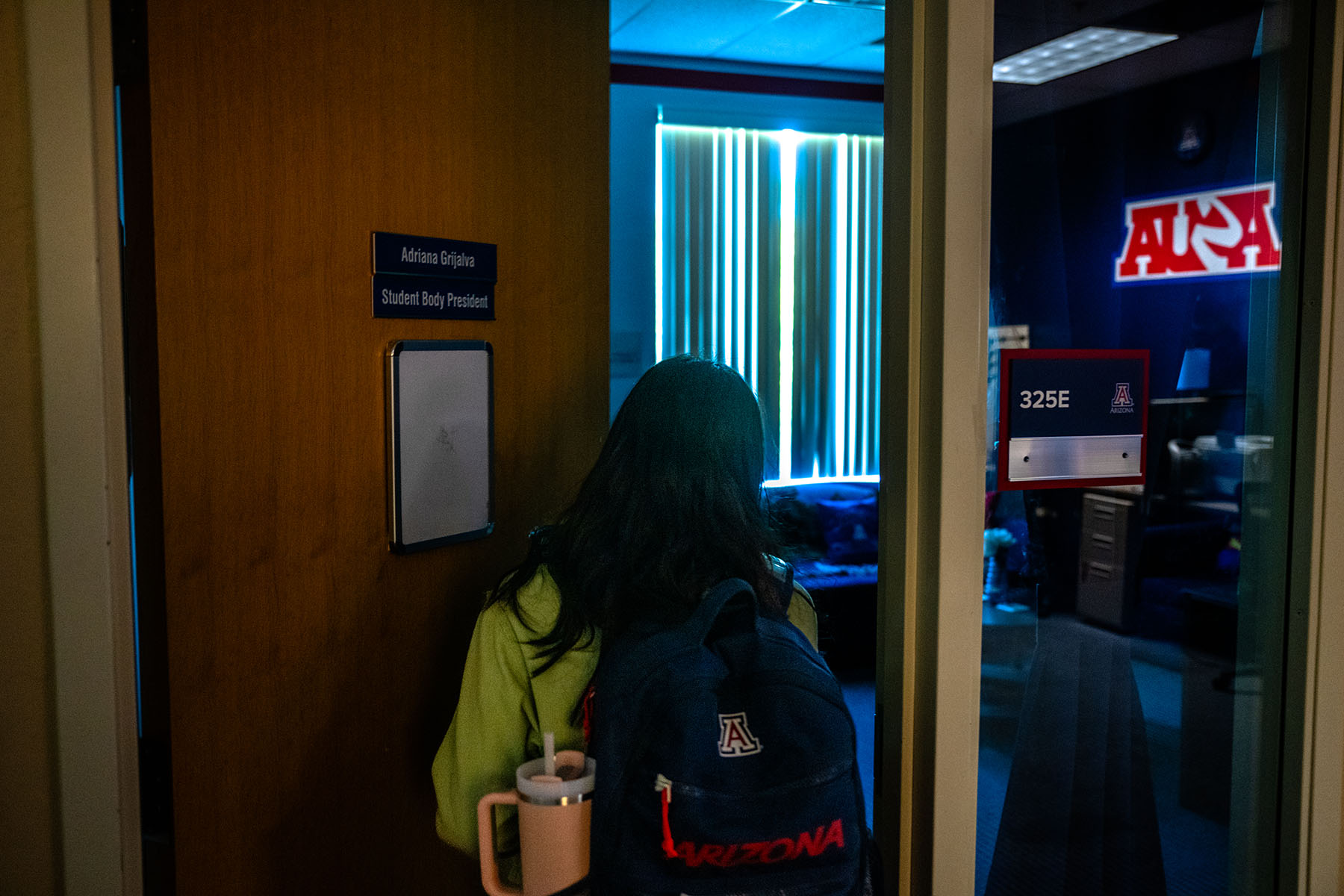 Adriana Grijalva stands in the doorway of her student body president office on the University of Arizona campus, facing away from the camera. The door displays her name and title, and the university's emblem is visible through a glass window.