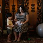 A pregnant woman sits on a chair, holding a baby doll, while her young child stands nearby, touching the doll. They are in a cozy corner of a house with toys and a yoga ball.