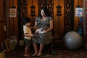 A pregnant woman sits on a chair, holding a baby doll, while her young child stands nearby, touching the doll. They are in a cozy corner of a house with toys and a yoga ball.