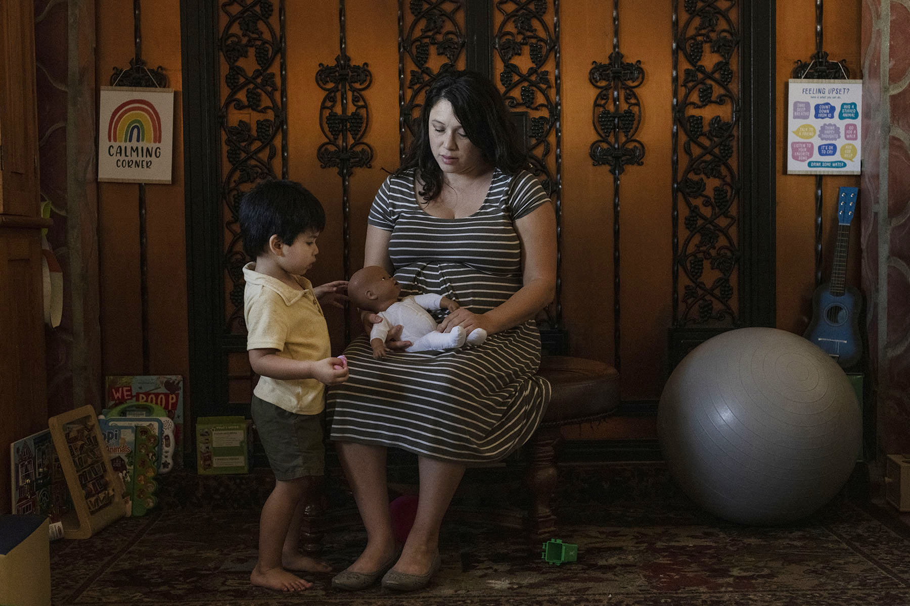 A pregnant woman sits on a chair, holding a baby doll, while her young child stands nearby, touching the doll. They are in a cozy corner of a house with toys and a yoga ball.