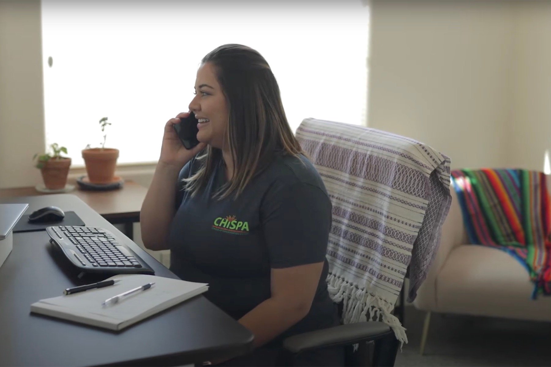 A woman is seen sitting at a desk smiling and speaking on the phone.