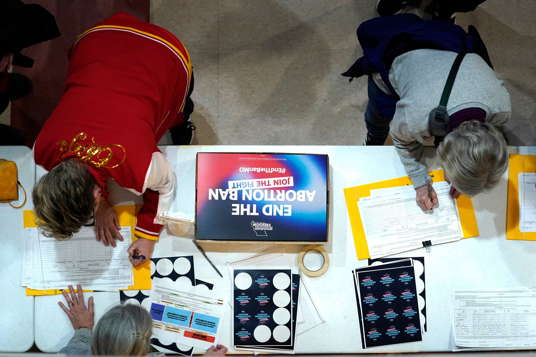 Two people sign petitions on a table, viewed from above.