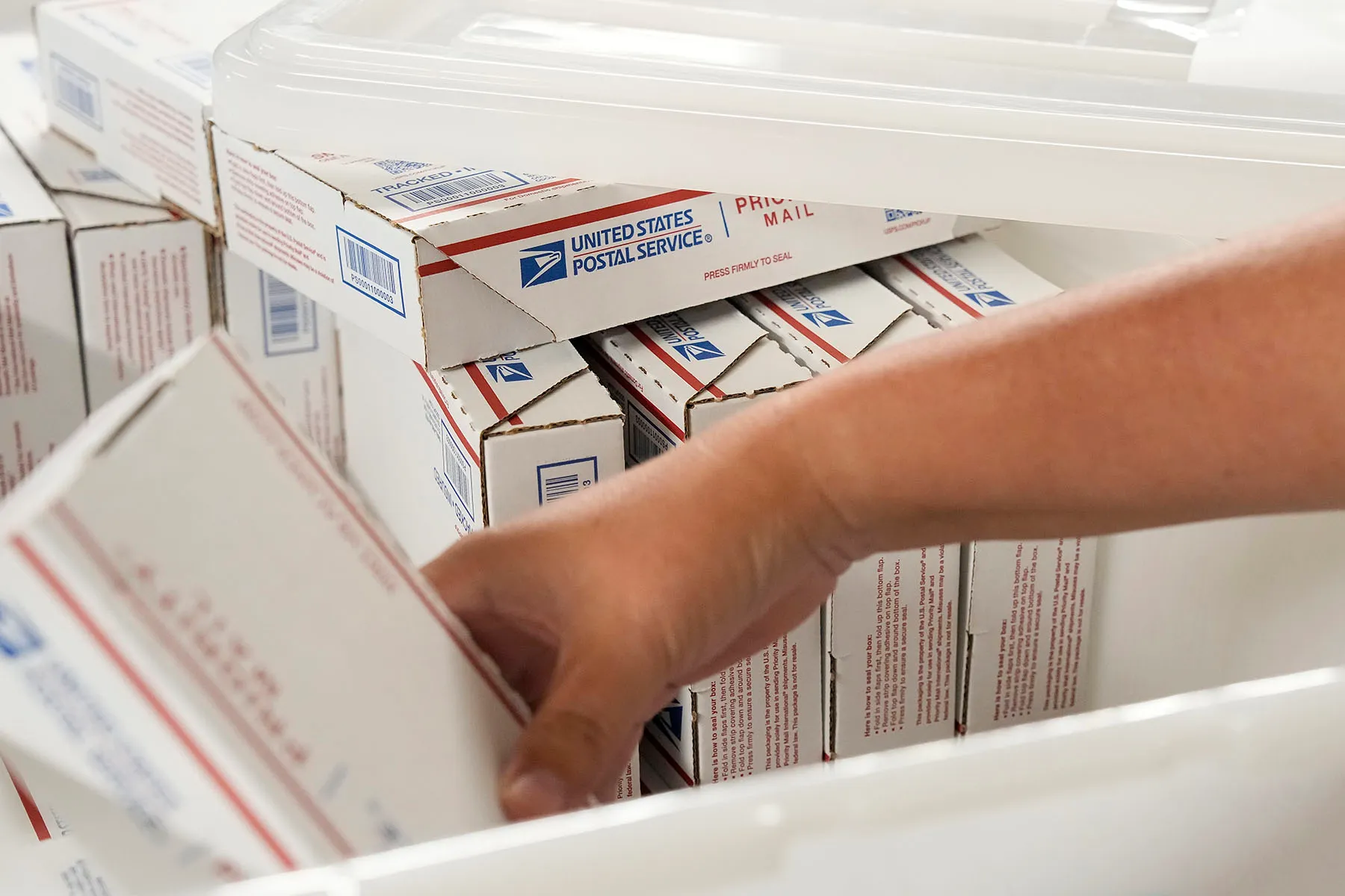 A close-up of a hand reaching for a box among many stacked United States Postal Service packages containing abortion medication.