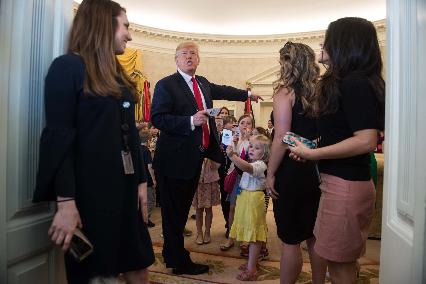 Several women and Donald Trump stand at the door watching a group of children in the Oval Office.