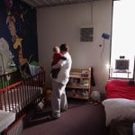 A mother holds an infant near a crib in a prison cell with a cartoon scene painted on the wall behind the crib.