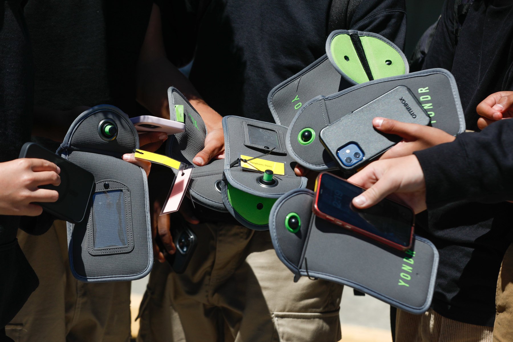 Varias manos jóvenes sostienen bolsas planas de color gris junto a teléfonos móviles mientras un grupo de estudiantes permanece unido.