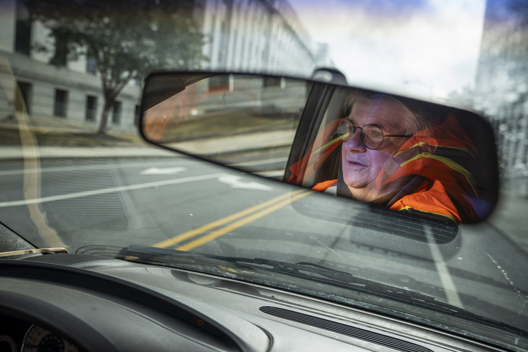 A rearview mirror reflects a driver and their orange vest framed by an empty city street.