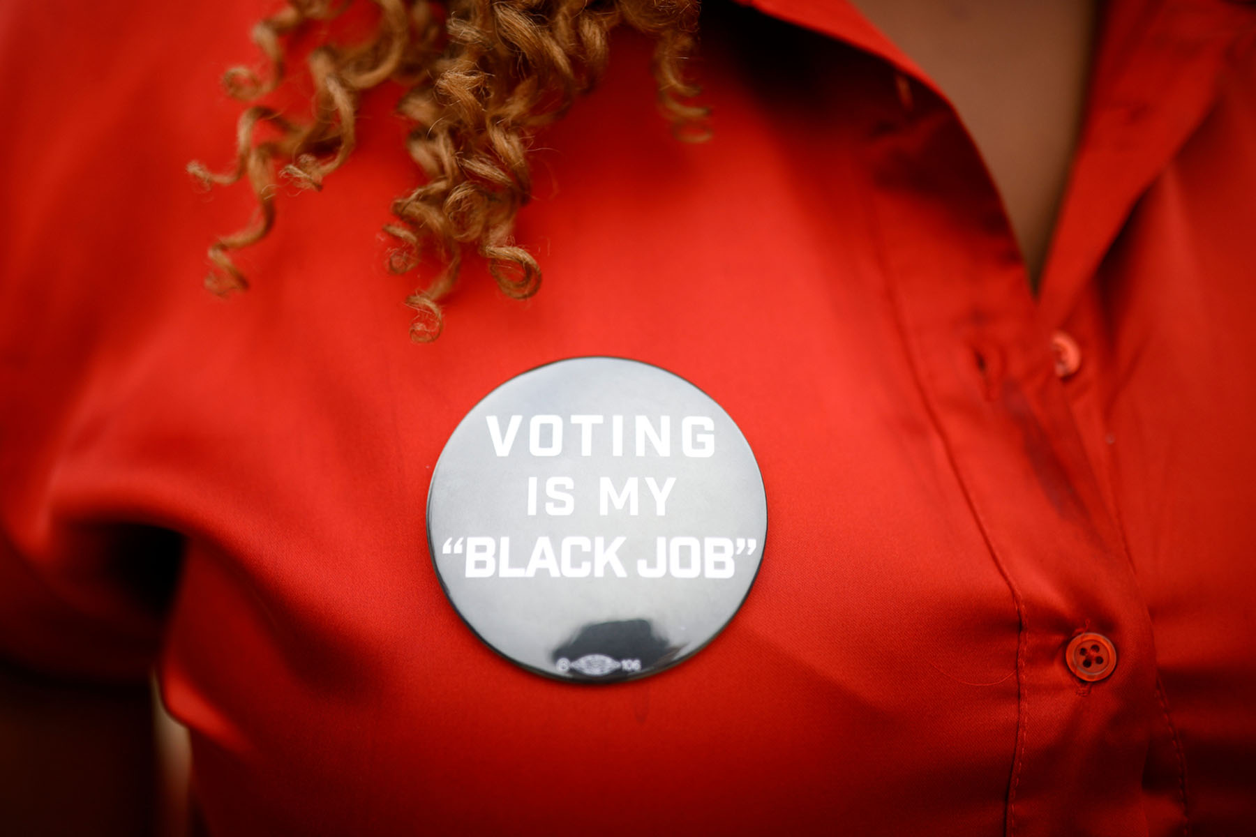 A button that says "voting is my black job" is pinned to a woman"s red shirt below her curly hair.