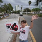 A woman stands waves at passing cars in the middle of the street while holding a sign that says 