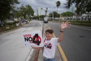 A woman stands waves at passing cars in the middle of the street while holding a sign that says 