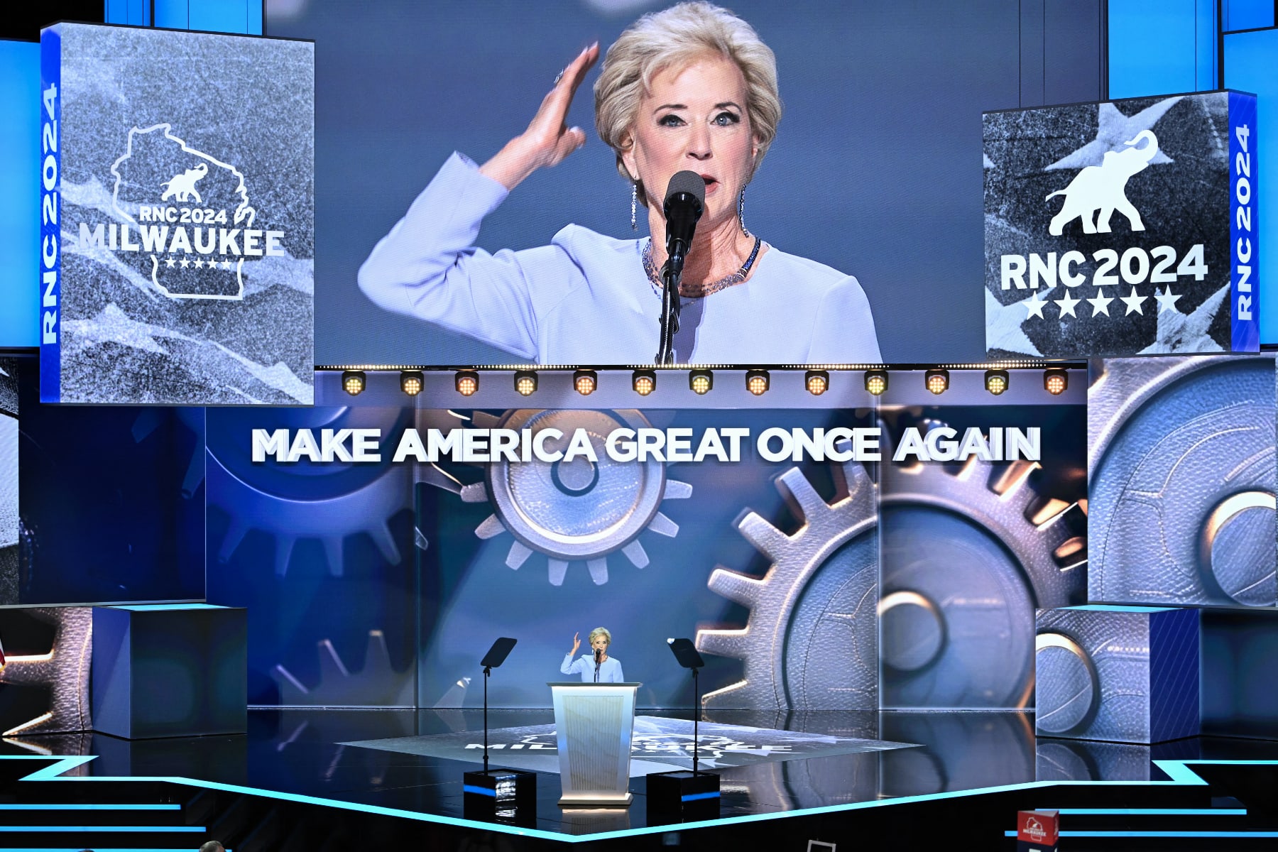 Linda McMahon speaks on stage in front of a screen with gears that says "Make America Great Once Again" and under a large screen showing her at the mic