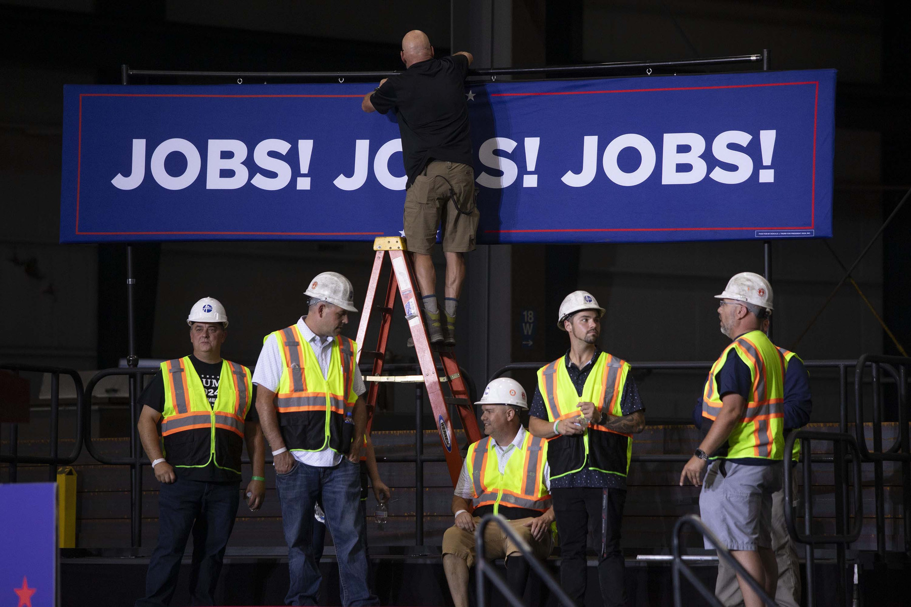 A man on a ladder hangs a "JOBS! JOBS! JOBS!" banner above a stage while other men in construction worker attire stand around.