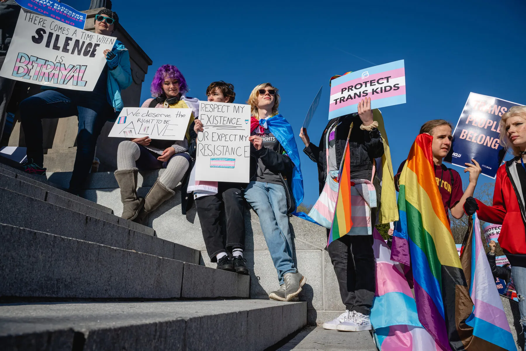 A line of young people stand along stairs, holding signs and LGBTQ+ flags during a rally.