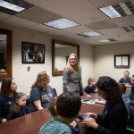 A group of parents and children from Little Lobbyists sit around a conference table during Rep. Jennifer Wexton’s first constituent meeting in 2019. Rep. Wexton stands at the table, smiling and engaging with attendees.