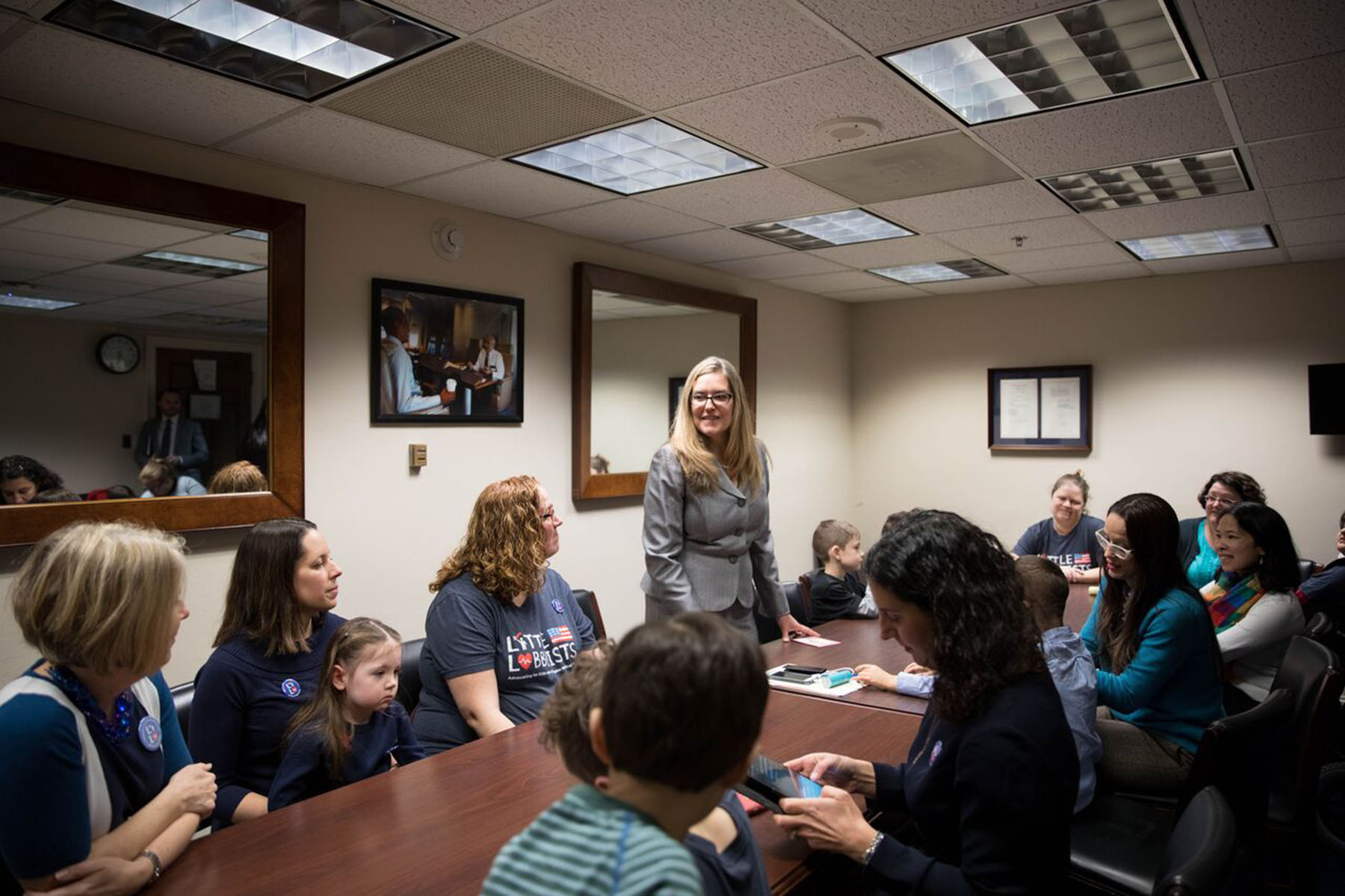 A group of parents and children from Little Lobbyists sit around a conference table during Rep. Jennifer Wexton’s first constituent meeting in 2019. Rep. Wexton stands at the table, smiling and engaging with attendees.