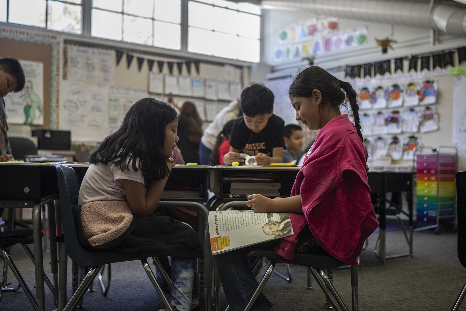 A close-up view of two young students as they sit in chairs facing each other. The student on the right wears a hot pink sweater while looking down at a workbook that rests on her thighs. The student on the left wears a light pink shirt and rest her hands on her chin.