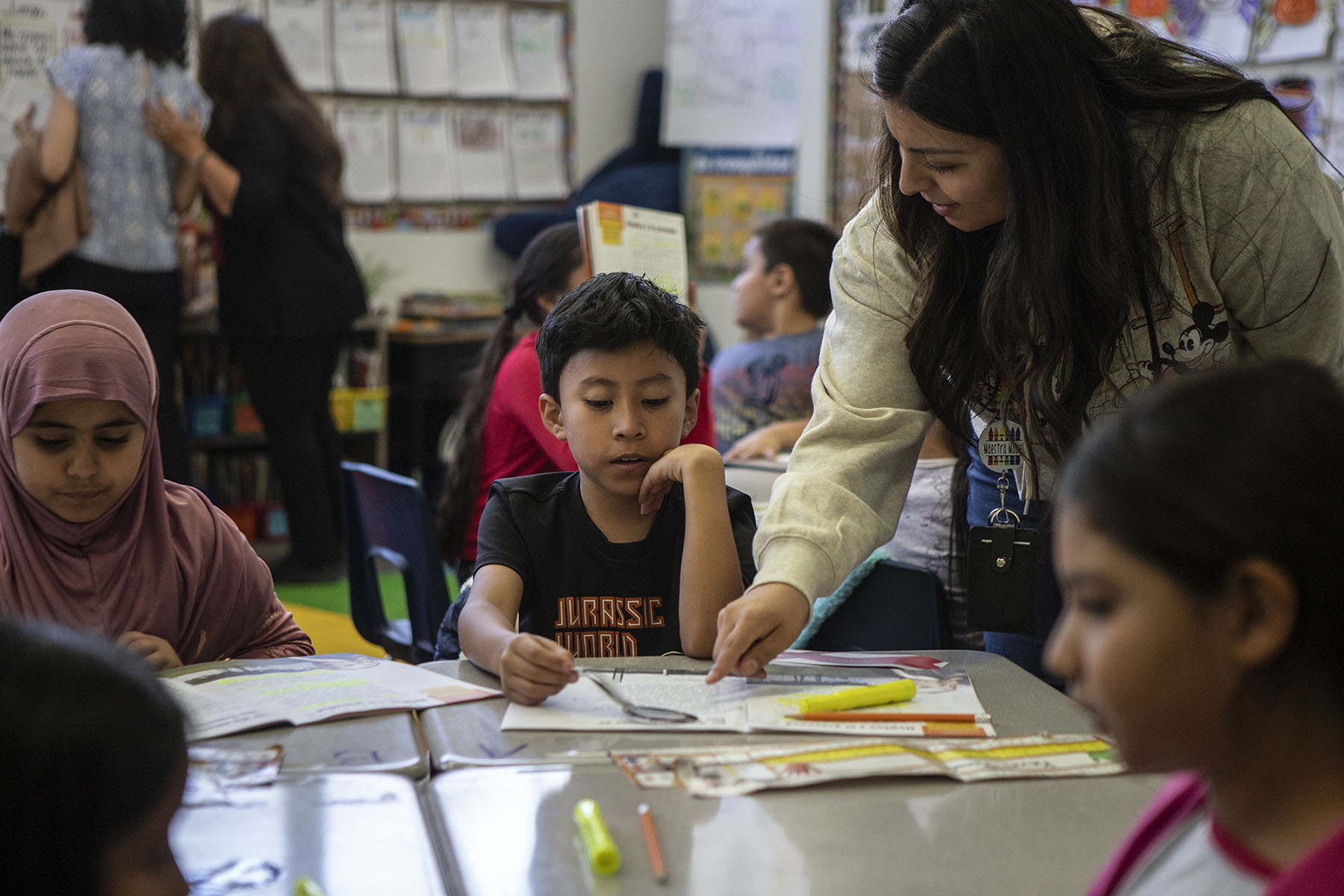 A teacher points at a book while a child sitting at their desk looks at it. Other students are visible in the frame. The setting is a classroom.