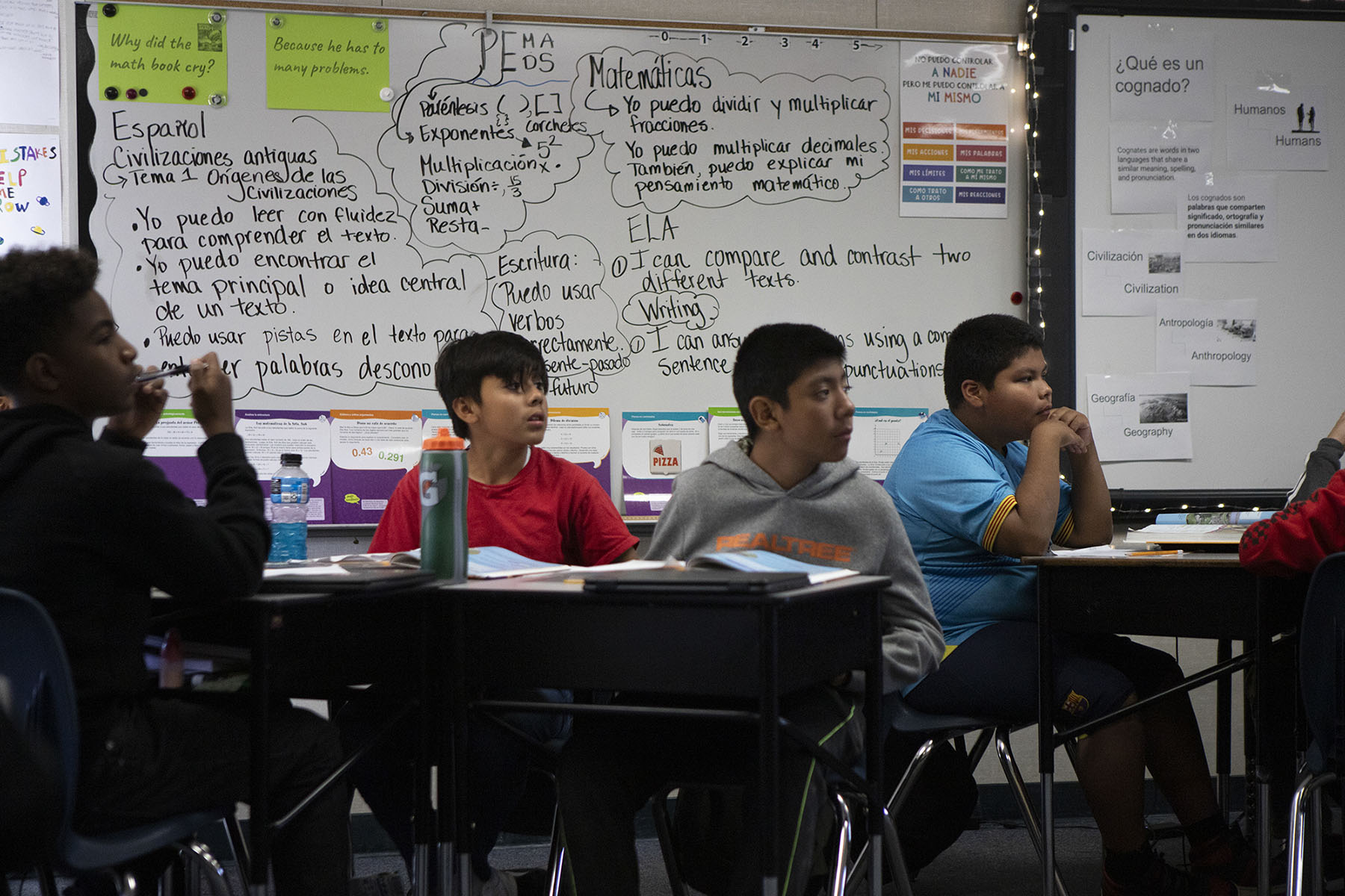 A row of students in a classroom sit at their desks listening to a lesson. A dry erase board with writing is visible behind them.