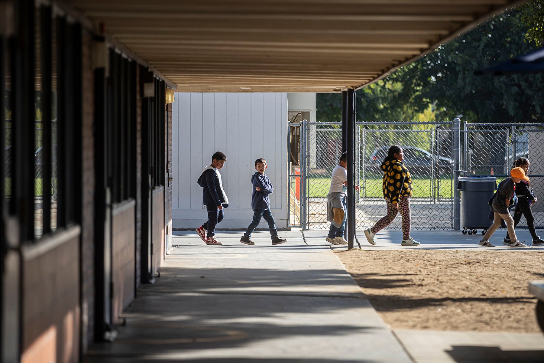 A line of children walk outdoors through a school campus. Fencing and other buildings are visible in the background.