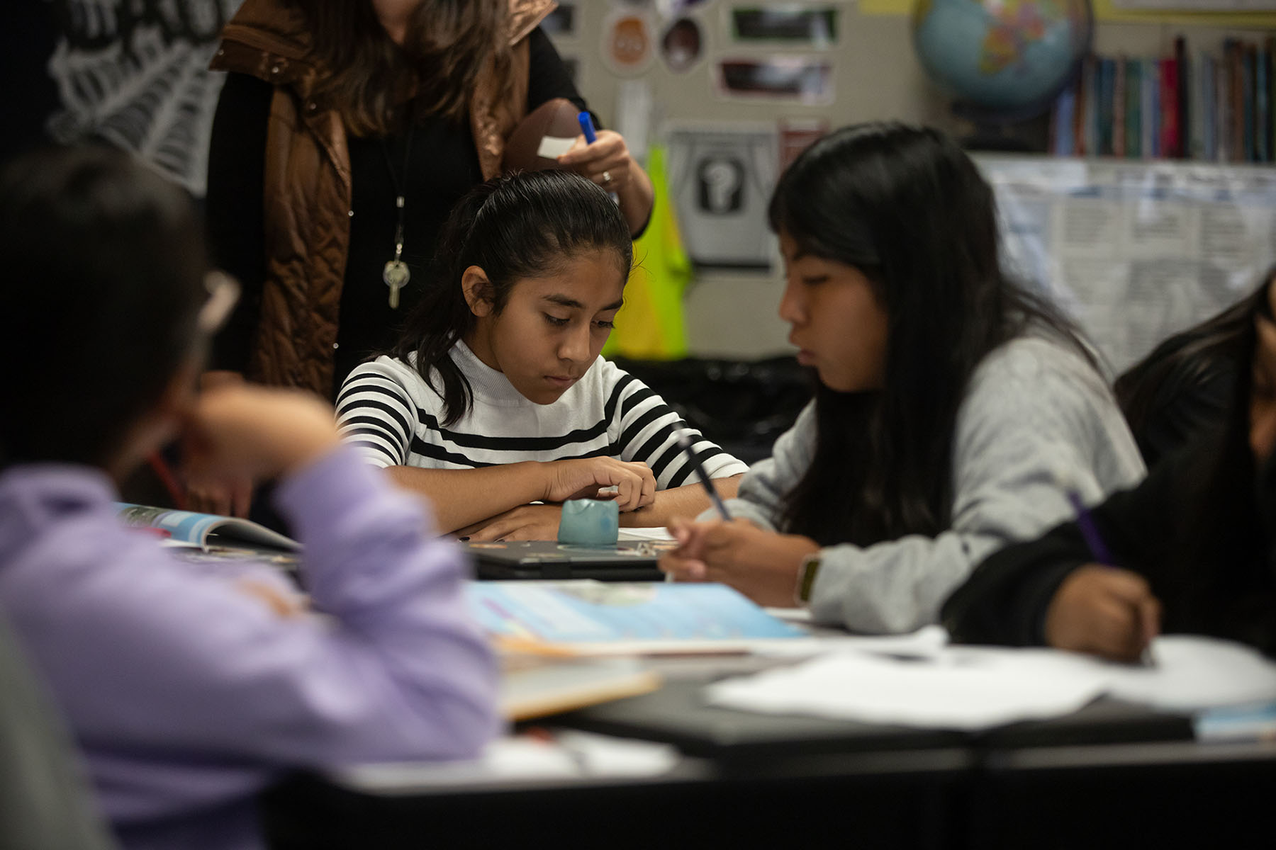 A student sits at their desk and reads their classwork while in class with other students. Students and teachers are visible and surround the student.