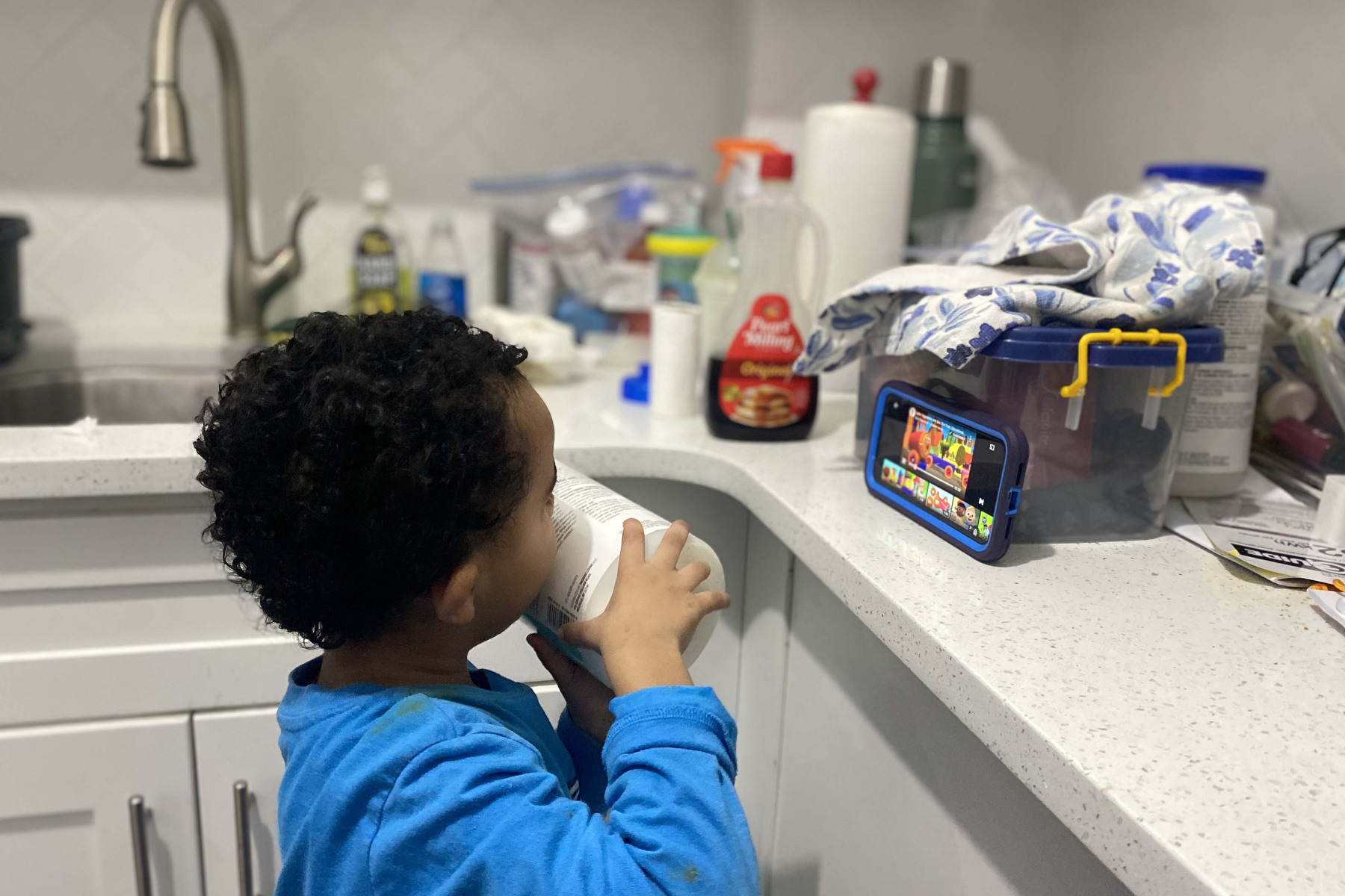 A young boy watches a cartoon on a cell phone in a kitchen.