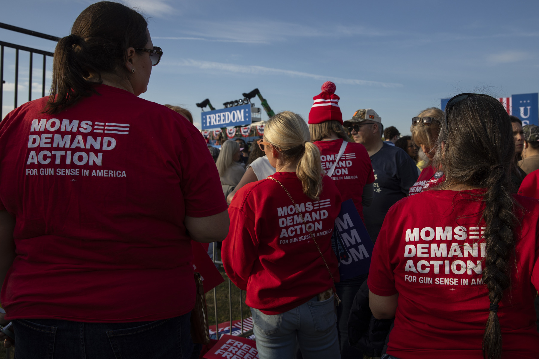 Several people wearing red shirts with the words "Moms Demand Action" face the stage at a political rally.