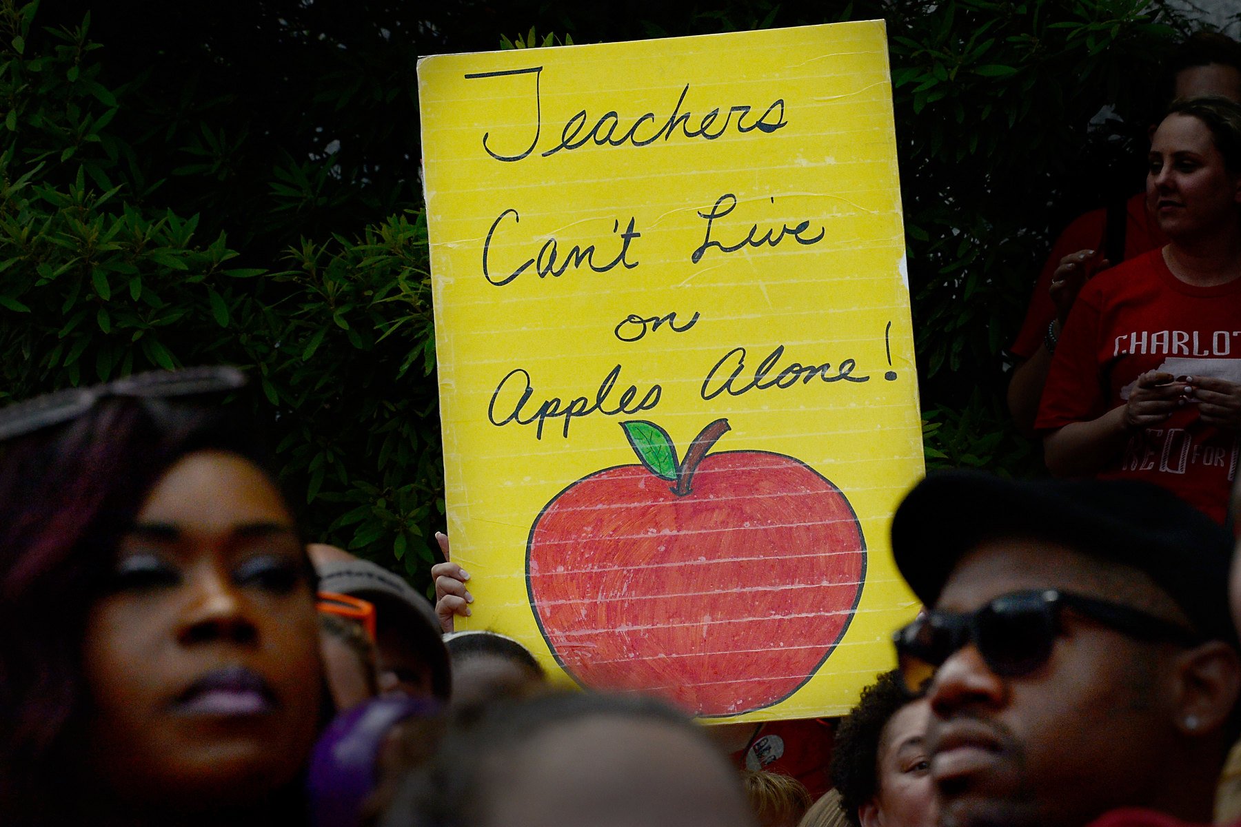 A group of people stand with a protest sign held up among them that reads “teachers can’t live on apples alone.”