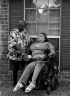 A woman holds the hand of another woman using a wheelchair in a portrait of the tow of them outside against a brick wall.