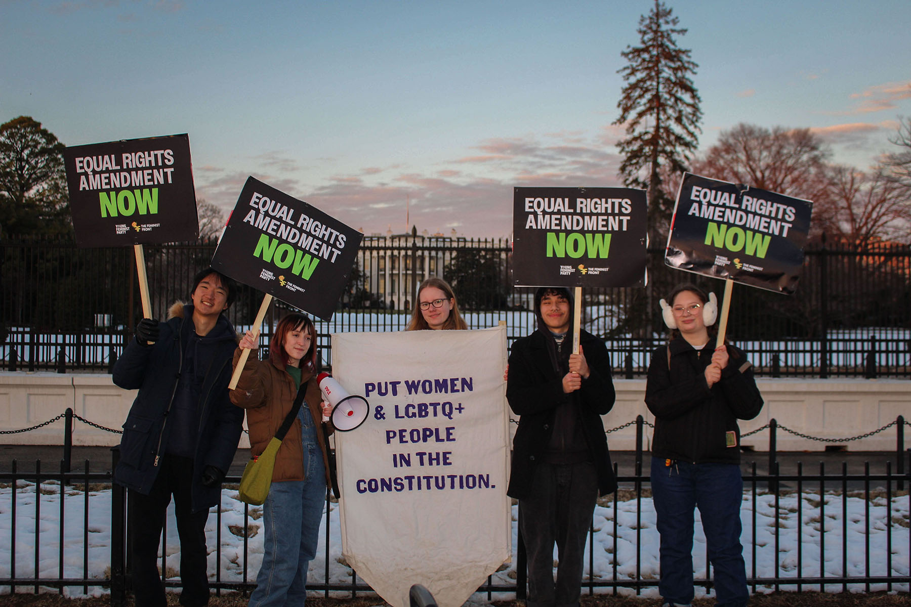 A group of young activists stand in front of the White House holding signs that read "Equal Rights Amendment NOW." One activist holds a large banner that says, "PUT WOMEN & LGBTQ+ PEOPLE IN THE CONSTITUTION." The ground is covered in snow.