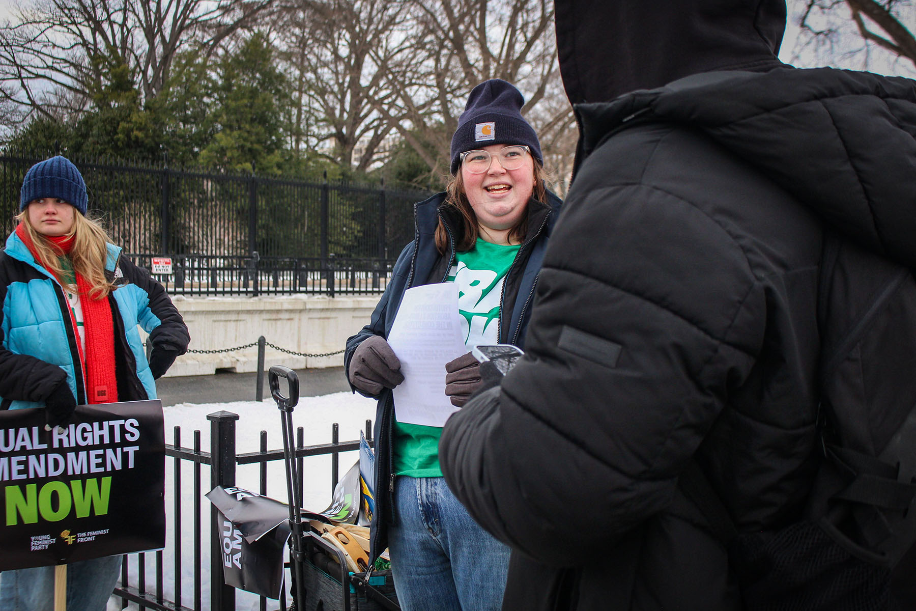 Rosie Couture dressed warmly in a blue coat and hat speaks to someone outside the White House, smiling while holding a piece of paper. Another person in a colorful scarf and jacket stands nearby, holding a sign supporting the Equal Rights Amendment.