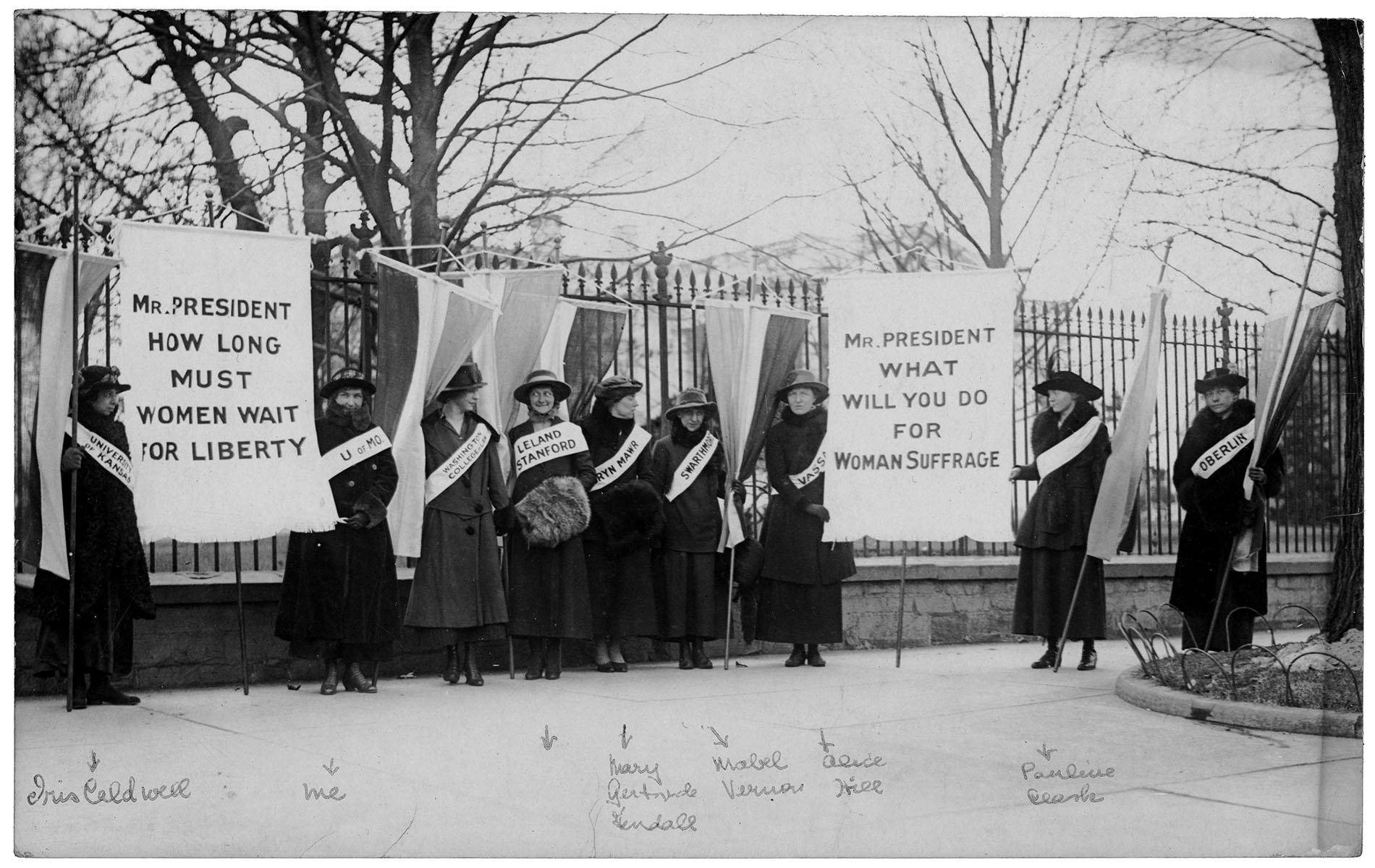 Suffragists picket with banners, wearing sashes that state the names of their colleges in January 1917.