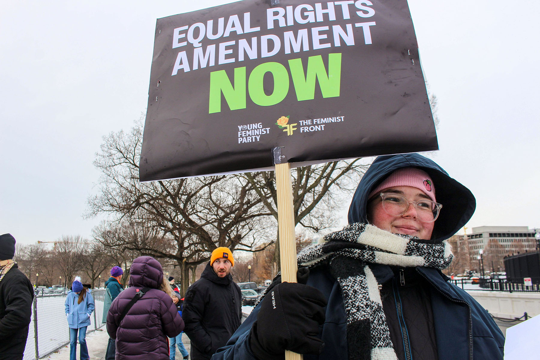 Rosie Couture, bundled in winter clothing with a scarf, hat, and gloves, smiles while holding a sign that says "Equal Rights Amendment NOW" in bold white and green letters. Other people and trees can be seen in the snowy background.