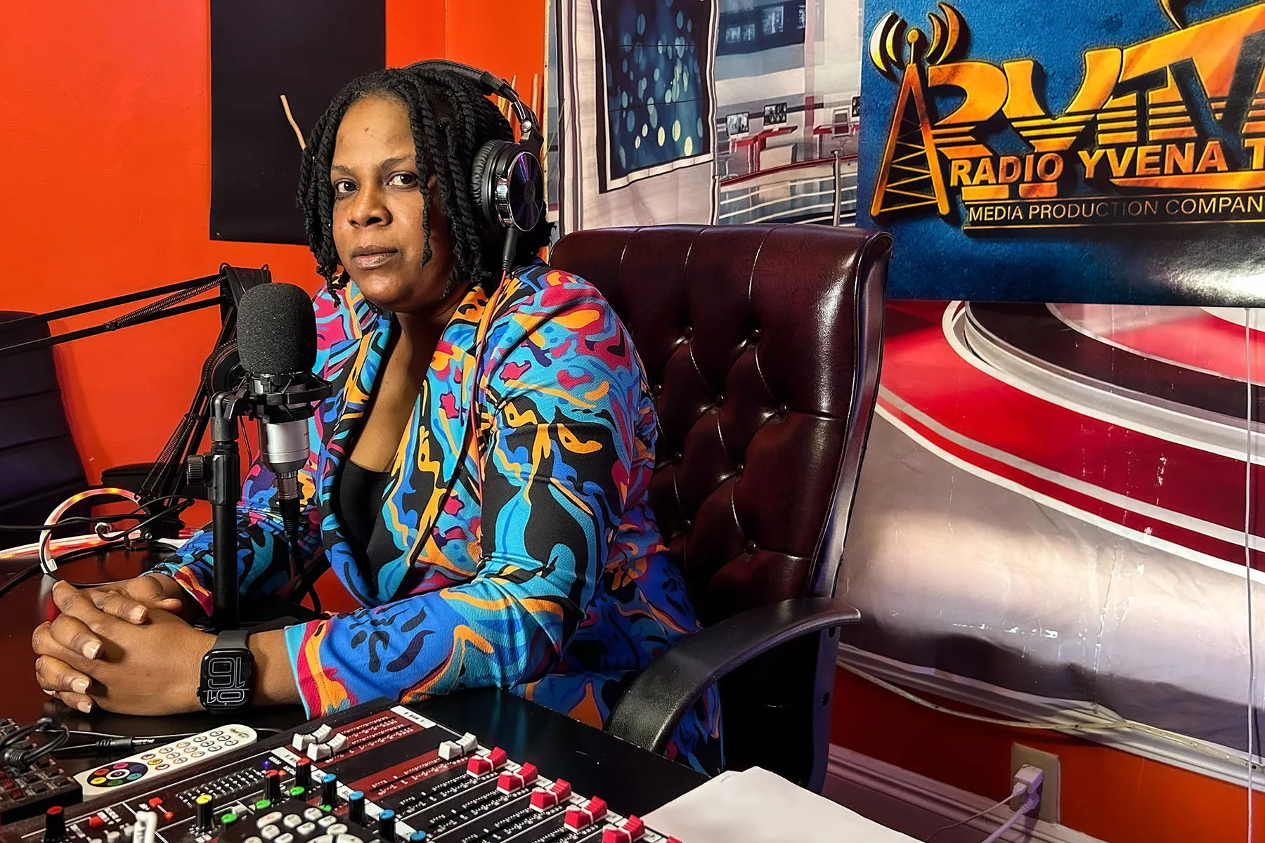 Yvena Jean François sits at her desk in her podcasting studio on Inauguration Day, January 20, 2024, in Springfield, Ohio. She wears a colorful patterned jacket and headphones while seated in front of a microphone and soundboard, with a vibrant studio backdrop featuring the logo of Radio Yvena TV.